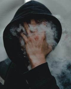Imagen de una persona con sudadera negra y capucha puesta, con la mano derecha en la cara, impidiendo que se pueda ver esta. Entre sus dedos sale mucho humo que se puede ver en toda la fotografía